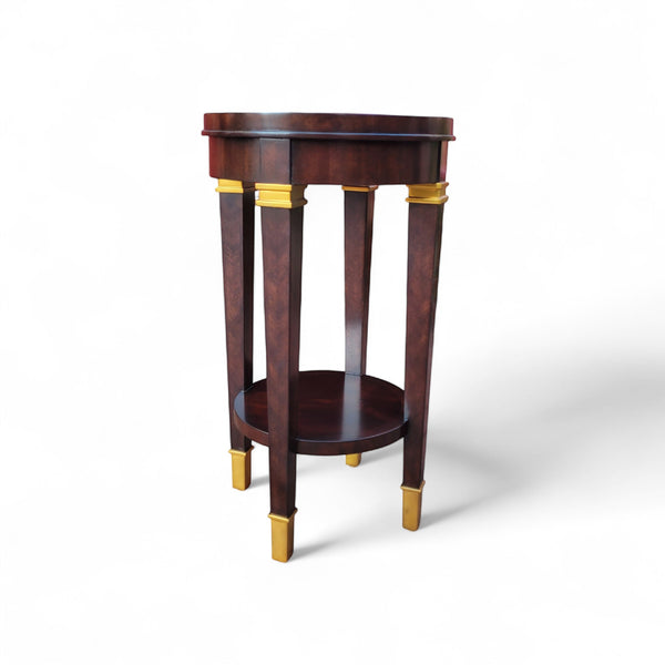 Tradition De France dark wood round accent table with gold accents on tapered legs and two tiers.